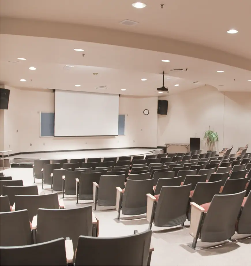 Empty lecture hall with rows of theater-style seating facing a projector screen and stage